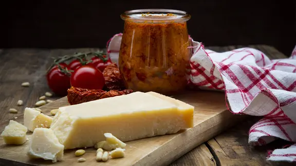 Parmesan, Tomaten und ein Glas mit Tomatenpesto stehen auf einem Holzbrett.