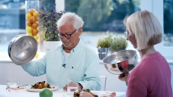 Doc Klasen und seine Patientin sitzen am gedeckten Tisch und lupfen die Deckel von den angerichteten Tellern.