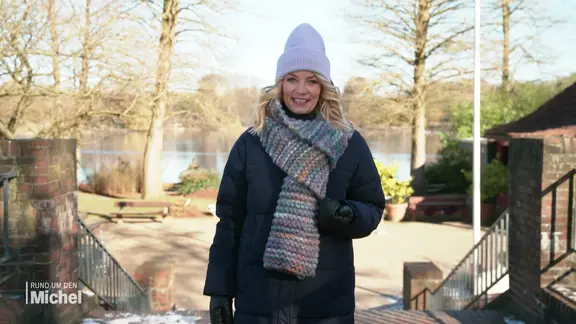 Moderatorin Dina Hille steht an einem Seeufer in einem Park.