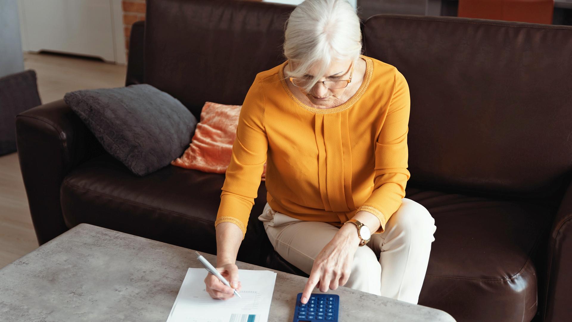 Eine ältere Frau sitzt an einem Wohnzimmertisch mit Stift, Papier und Taschenrechner | colourbox, Piotr Adamowicz