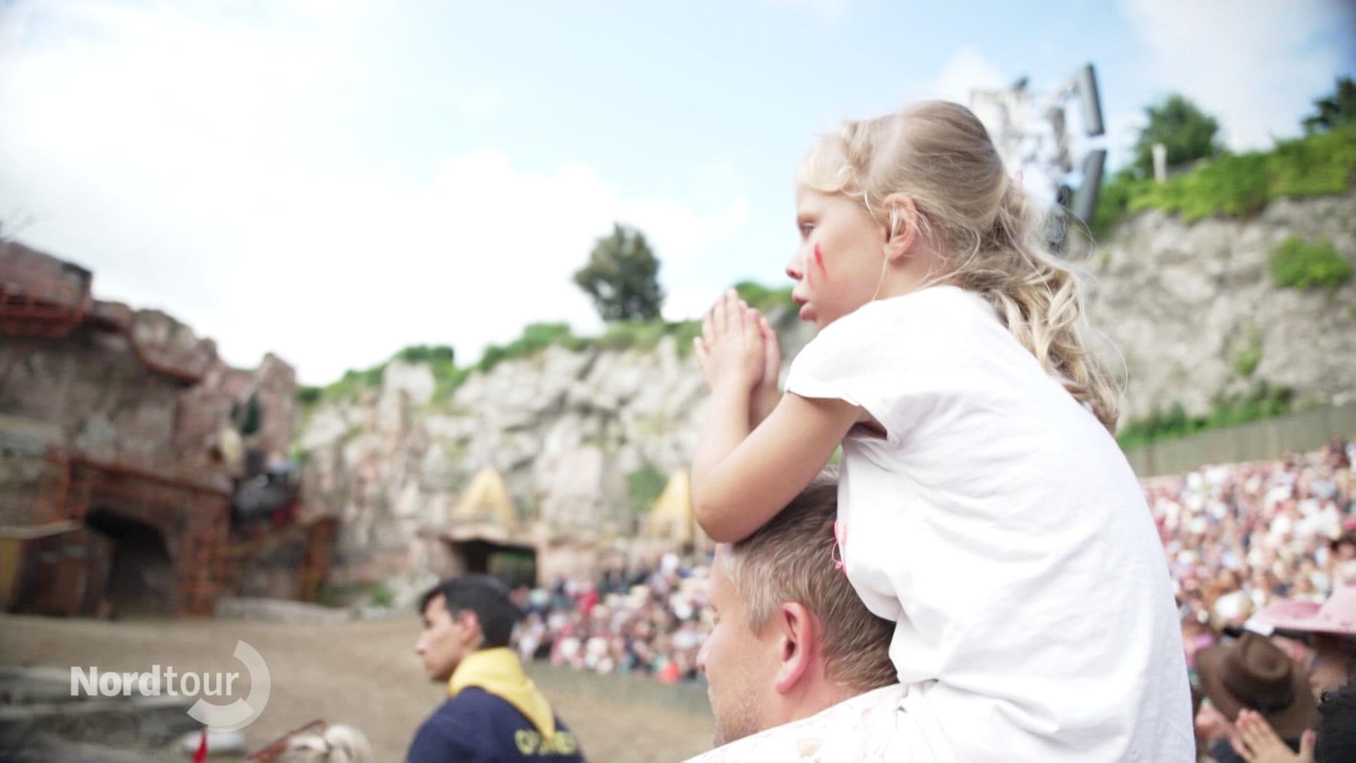 Ein kleineres Mädchen sitzt auf den Schulter eines Mannes und schaut bei den Karl-May-Festspielen zu. | Screenshot