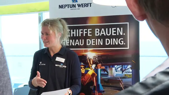 Ein Mitarbeiter der Neptun Werft informiert Messebesucher. | Screenshot Ein Mitarbeiter der Neptun Werft informiert Messebesucher.