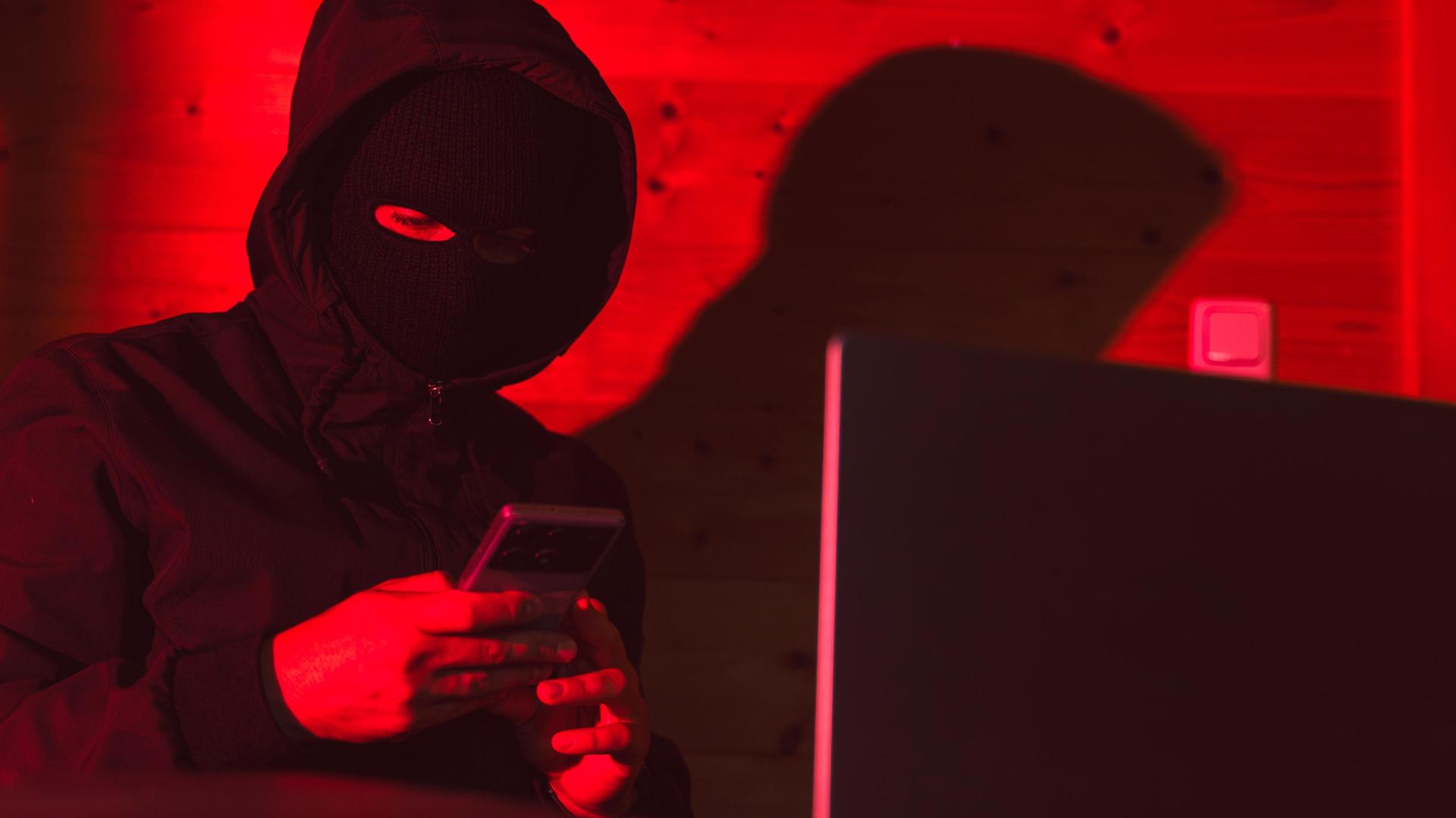 Ein maskierter Täter mit Handy und Laptop. (Themenbild) | CHROMORANGE, Michael Bihlmayer