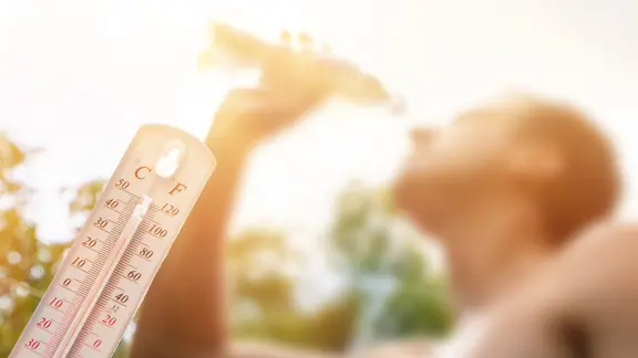 Eine Mann mit freiem Oberkörper trinkt aus einer Flasche. Im Vordergrund ein Thermometer, das mehr als 30 Grad anzeigt.