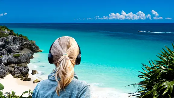 Eine Frau mit Kopfhörern blickt auf das Meer mit idyllischer Küstenlandschaft.