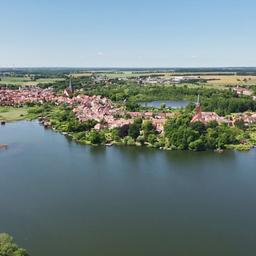 Eine kleine Stadt mit roten Dächern und mehreren Kirchtürmen liegt auf einer Halbinsel am Ufer eines großen Sees, umgeben von grünen Bäumen und Feldern unter klarem Himmel.