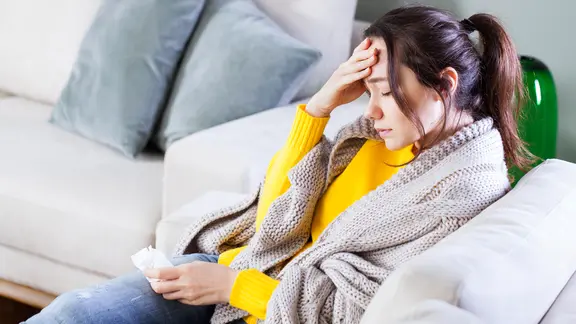 Eine kranke Frau sitzt auf einem Sofa und fasst sich an die Stirn. | Colourbox, Ahmet Eylem Misirligül Eine kranke Frau sitzt auf einem Sofa und fasst sich an die Stirn.