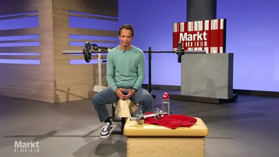 Moderator Jo Hiller im Studio von "Markt": Er sitzt auf einer Handtelbank, vor ihm ein Sprungkasten mit Handtuch, Wasserflasche und einer Hantel.