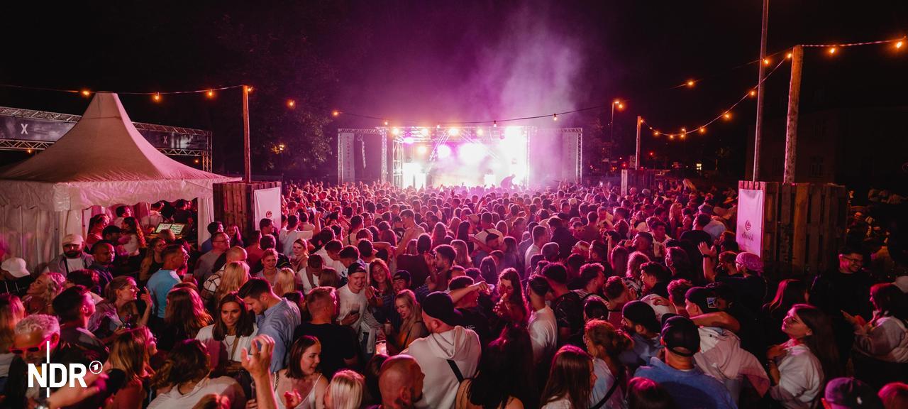 Eine große Menschenmenge steht nachts vor einer hell erleuchteten Bühne. Auf der Bühne findet ein Konzert statt. | EXC Media Eine große Menschenmenge steht nachts vor einer hell erleuchteten Bühne. Auf der Bühne findet ein Konzert statt.