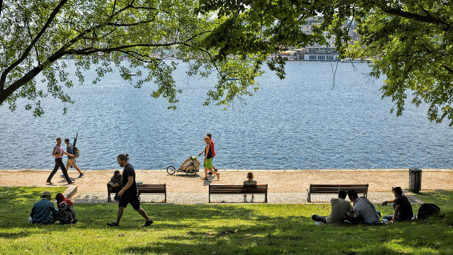 Menschen gehen bei Sommerwetter an der Alster in Hamburg entlang, andere liegen unter Bäumen im Schatten. | IMAGO Images / Zoonar