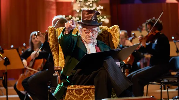 Die Weihnachtsgeschichte "A Christmas Carol" von Charles Dickens wird vorgelesen von einem alten Mann mit Zylinder im Sessel sitzend, im Hintergrund ein leuchtender Weihnachtsbaum und ein Orchester