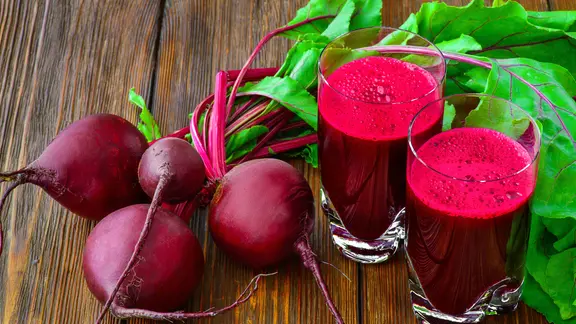 Frische Rote-Bete-Knollen mit Blättern und zwei Gläser mit Rote-Bete-Saft auf einem Holztisch.