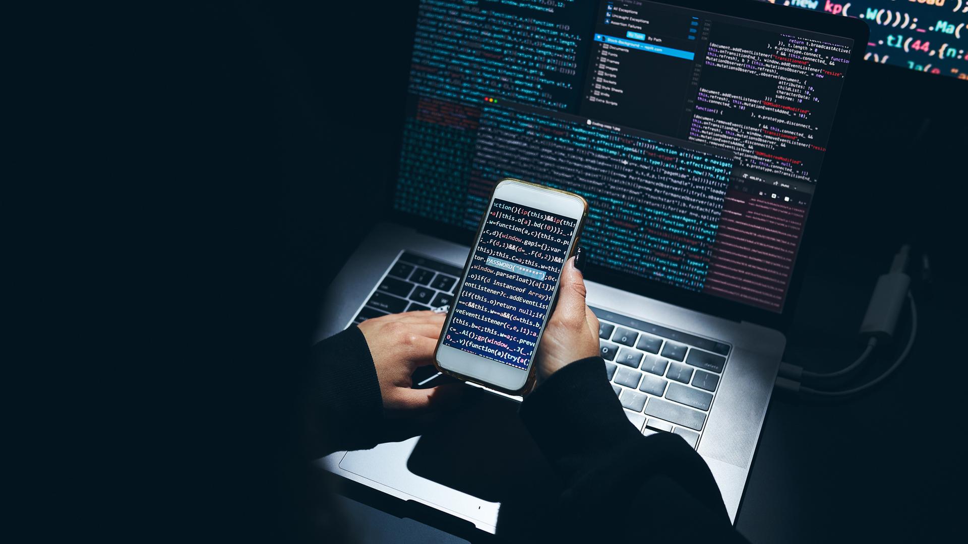 Hände halten ein Smartphone vor einem Laptop. Auf den Bildschirmen befinden sich abstrakte Codes. | photocase.de, przemekklos