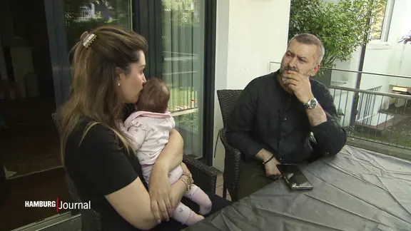 Eine Frau mit Baby und ein Mann, der besorgt schaut, sitzen zusammen am Tisch.