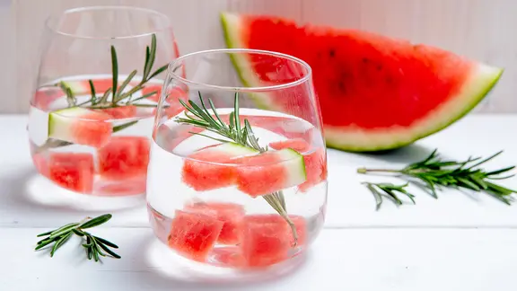 Zwei Gläser Wasser mit Melonenstückchen und Rosmarin stehen auf einem Tisch.