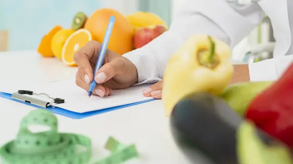 Stoffwechseldiät, Symbolbild: Person mit Arztkittel schreibt auf eine Kladde, daneben liegen ein Maßband, Obst und Gemüse.