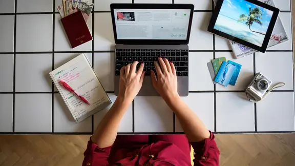 Eine Frau tippt an einem Laptop. Neben ihr liegen ein Notizblock, ein Reisepass, ausländisches Geld, Kreditkarten, ein Fotoapparat und ein Tablet mit dem Bild einer Palme.