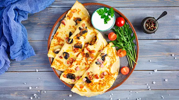 Ein Pfannkuchen mit Schinken und Pilzen sowie Tomaten und Rucola auf einem Teller.