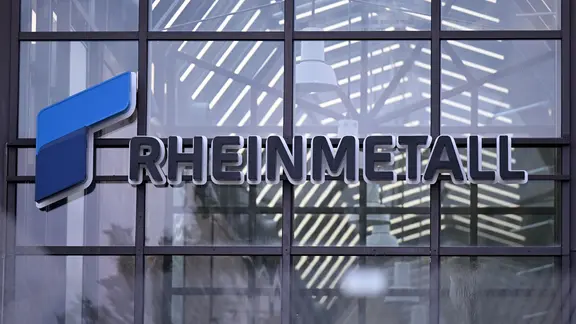 Das Logo des Rüstungskonzerns Rheinmetall ist hinter einem Zaun (verschwommen im Vordergrund) am Eingang eines Rheinmetall-Werks zu erkennen. 