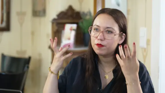 Ronya Othmann sitzt in einem hellen Raum mit Holzwänden, trägt eine dunkle Bluse und große Brille. Sie gestikuliert mit beiden Händen, spricht konzentriert und wirkt nachdenklich.