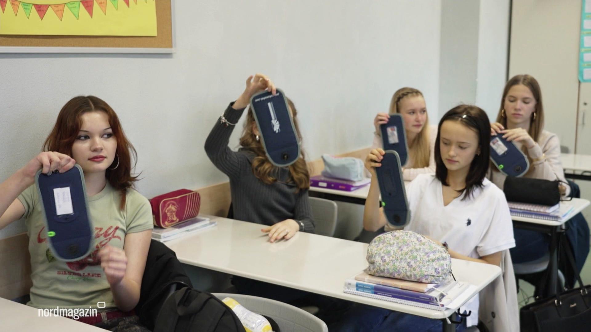 Schülerinnen sitzen hintereinander in der Klasse und zeigen ihre Handytaschen. | Screenshot