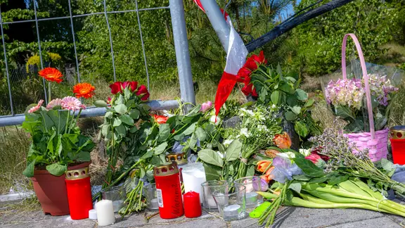 Auf dem Bild sind Kerzen, Blumen und ein Absperrband der Polizei an einem Weg zu sehen. An der Stelle soll ein Mann seine Ehefrau absichtlich überfahren haben.