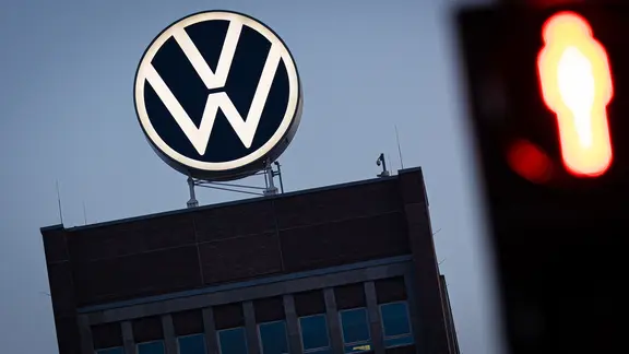 Das VW-Logo leuchtet auf dem Dach des Markenhochhauses im Volkswagen Stammwerk am frühen Morgen hinter einer rot zeigenden Fußgänger-Ampel.