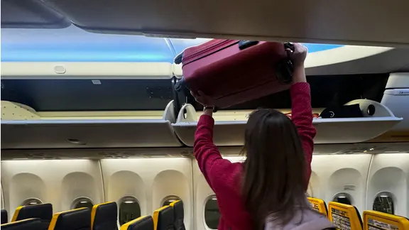 Eine Frau hebt ihr Handgepäckskoffer in die Gepäckablage eines Flugzeugs. 