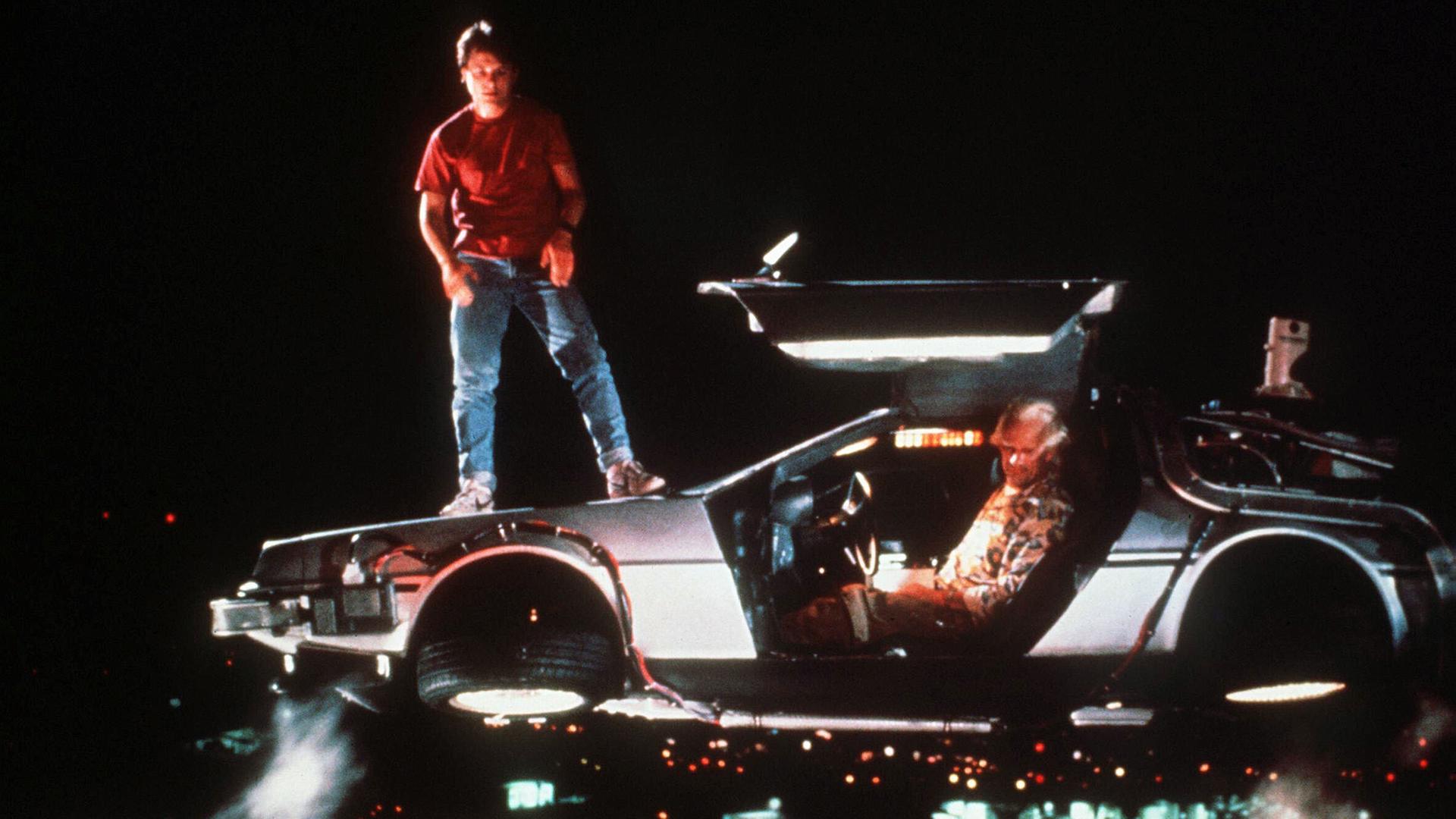 Michael J. Fox steht auf dem Dach eines schwebenden DeLorean DMC-12, dessen Türen geöffnet sind. Christoph Lloyd sitzt am Steuer des futuristisch umgebauten Sportwagens. | picture alliance/United Archives