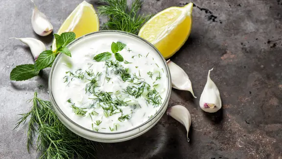 Ein Schälchen mit Joghurtdip, daneben liegen Kräuter, Knoblauchzehen und Zitronenspalten