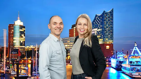 Ole Wackermann und Maiken Nielsen stehen vor einem Hintergrund, der den Hamburger Hafen und die Elphilharmonie am Abend zeigt, und lächeln in die Kamera.