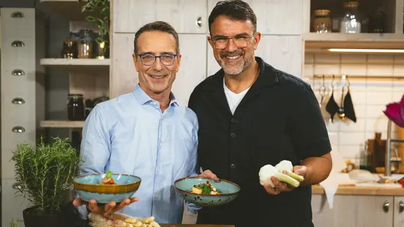 Tarik Rose und Ernährungs-Doc Matthias Riedl halten zwei Teller mit Gemüsegerichten in die Kamera.