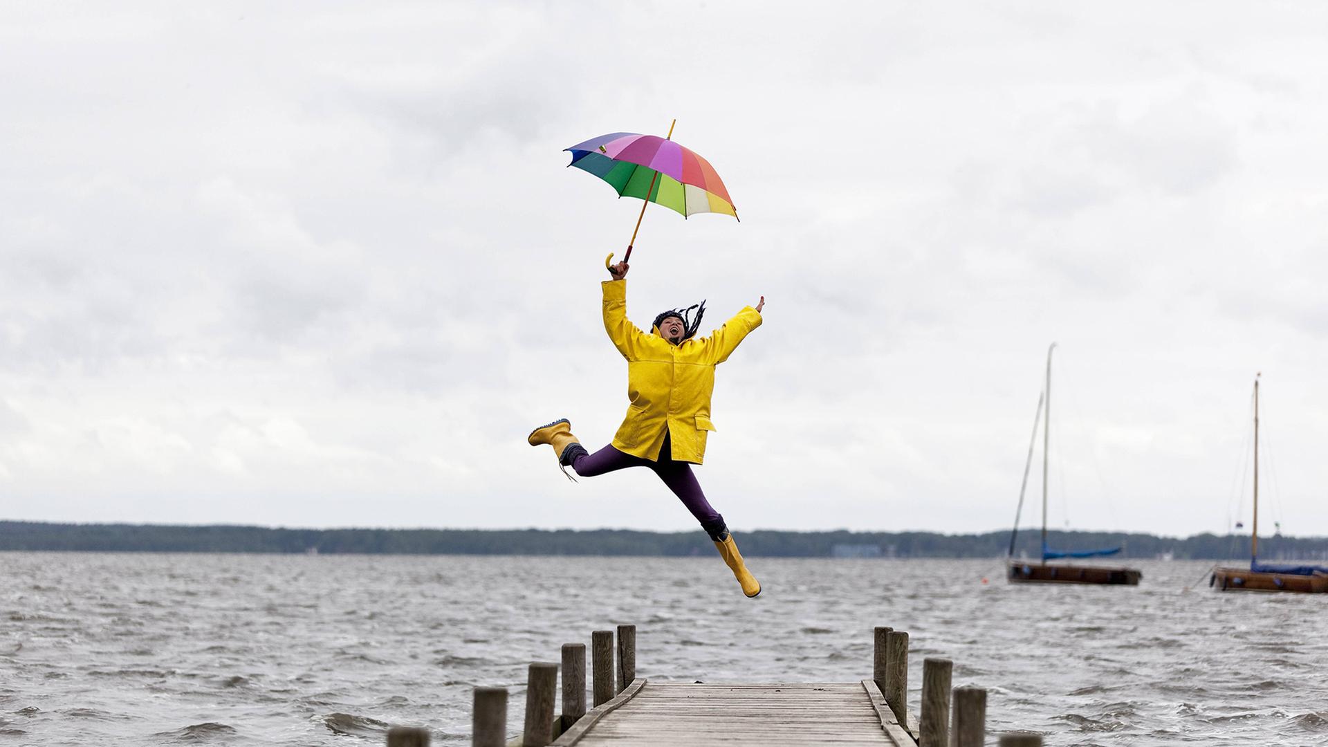 Eine Frau in Regenjacke und mit Regenschirm springt auf einem Steg am Wasser | NDR, Westend61