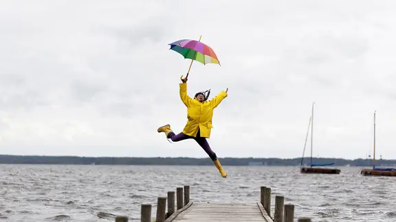 Eine Frau in Regenjacke und mit Regenschirm springt auf einem Steg am Wasser