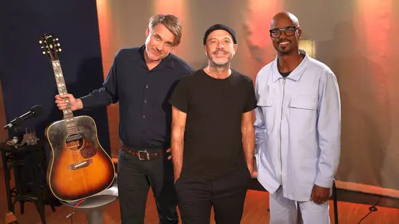 Drei Männer stehen nebeneinander, der Mann ganz links hält eine Gitarre in der Hand.