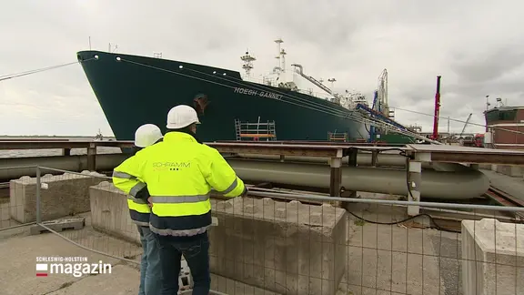 Zwei Männer in Sicherheitskleidung stehen vor einem Schiff.