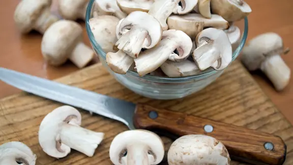 Aufgeschnittene Champignons in einer Glasschale und auf einem Holzbrett mit Messer