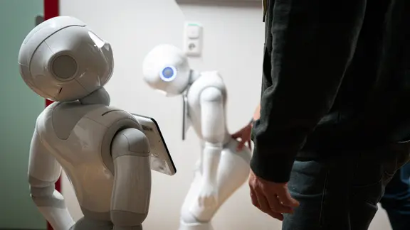 Roboter Pepper befindet sich in einem Flur des Instituts für Informationssystem in Lübeck und schaut hoch. Im Hintergrund wird ein zweiter Roboter seiner Art durch den Gang geschoben.