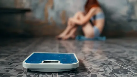 Eine Personenwaage im Vordergrund, dahinter unscharf ein sehr dünnes junges Mädchen.
