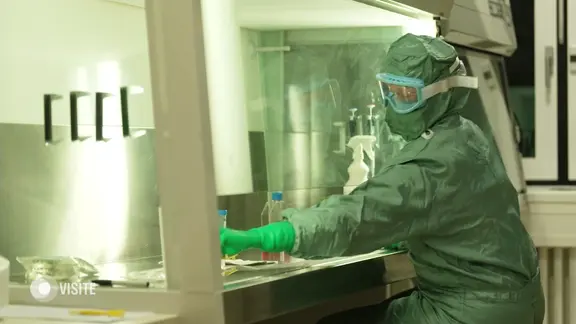 Eine Person im grünen Ganzkörperschutzanzu sitzt an einem beleuchteten Labortisch.