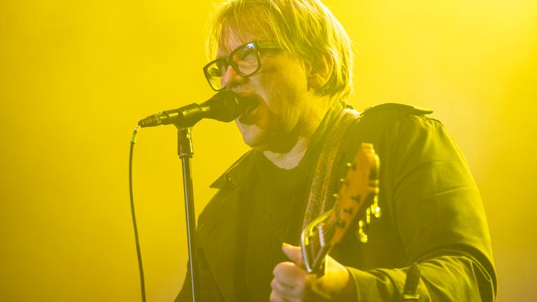 Sänger Sven Regener von Element of Crime steht auf einer Bühne in gelbem Licht, singt ins Mikrofon und spielt Gitarre. Er trägt eine Brille und eine dunkle Jacke.
