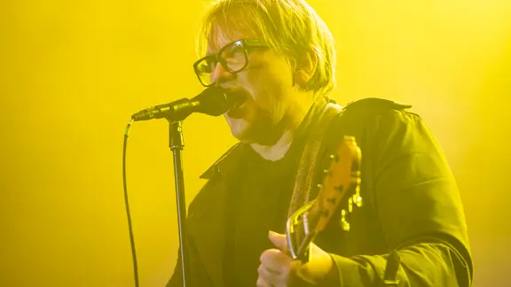 Sänger Sven Regener von Element of Crime steht auf einer Bühne in gelbem Licht, singt ins Mikrofon und spielt Gitarre. Er trägt eine Brille und eine dunkle Jacke.