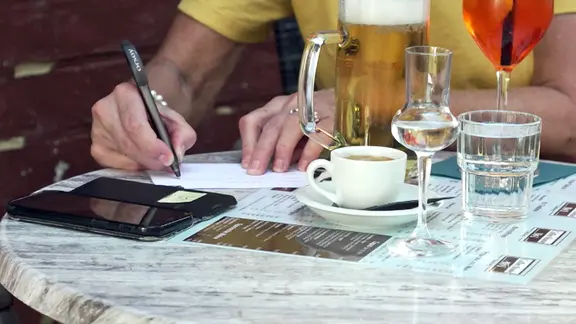 Person füllt einen Zettel aus, auf dem Tisch stehen Kaffee und Bier.