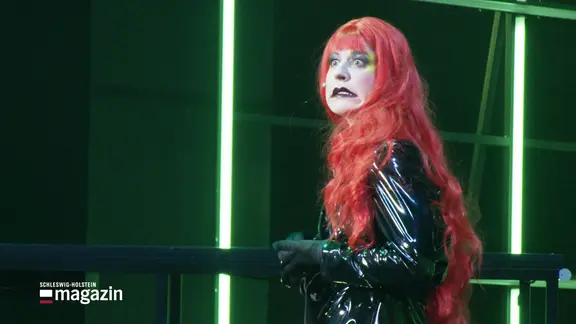 Eine Frau mit roten Haaren und weiß geschmicktem Gesicht auf einer Bühne.