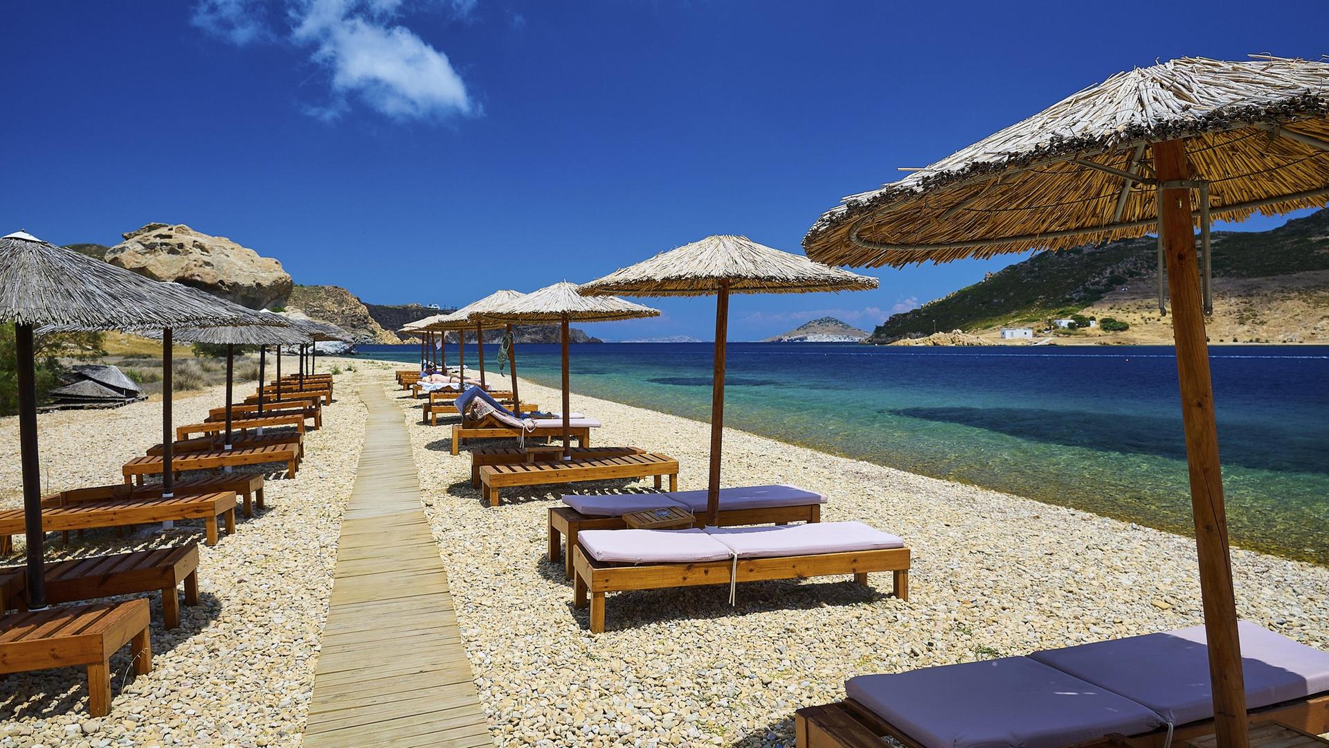 Liegestühle und Sonnenschirme stehen an einem am Strand mit Holzweg mit klarer  Sicht auf das Meer und den blauen Himmel. | picture alliance / imageBROKER | Ralf Adler