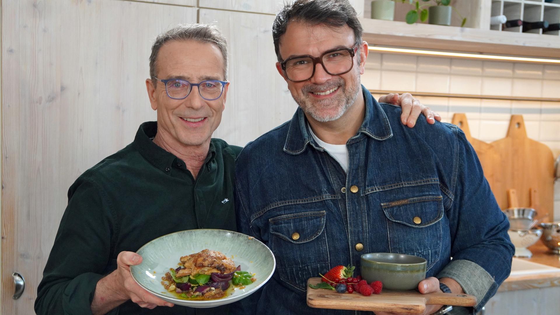 Tarik Rose und Ernährungs-Doc Matthias Riedl halten einen Teller mit knusprigem Huhn und eine Schale mit Creme brûlée in die Kamera. | NDR, Florian Kruck