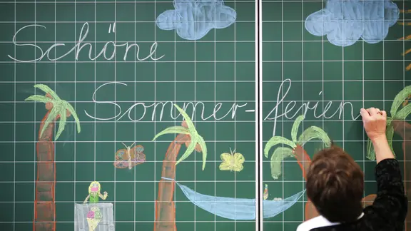 Eine Lehrerin schreibt "Schöne Sommerferien" an die Tafel