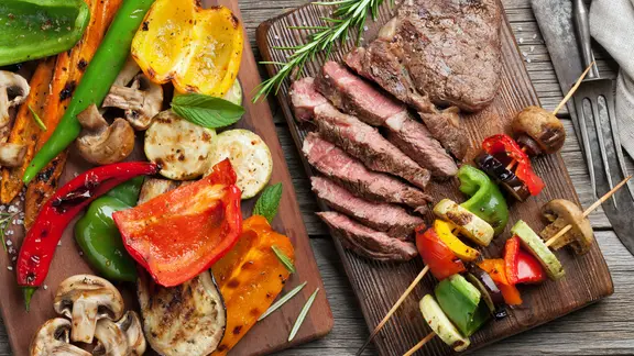 Auf Holzbrettern liegen gegrillte Gemüsestücke, zwei gegrillte Gemüsespieße und ein Stück angeschnittenes Rindfleisch.