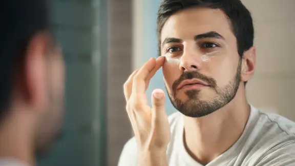 Ein junger Mann pflegt die Haut unter den Augen mit einer Creme.