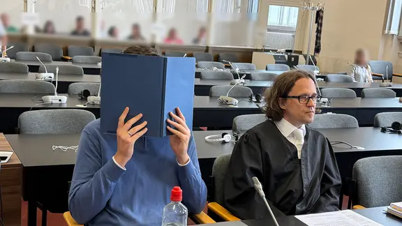In einem Gerichtssaal sitzt ein Mann neben seinem Verteidiger. Der Mann hält ein aufgeklapptes Buch vor seinen Kopf, um nicht erkannt zu werden